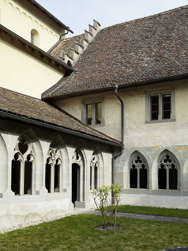 Vue de la cour intérieure du cloître. Au centre, il y a l’angle nord-est du cloître. Au premier plan, on voit la pelouse et un rosier sans fleurs. À gauche, il y a l’aile nord du cloître avec la façade sud de l’église. À droite, il y a l’aile est du cloître. Au deuxième étage, on voit deux fenêtres du dortoir. Il fait jour, le ciel est un peu couvert. La cour est ensoleillée.